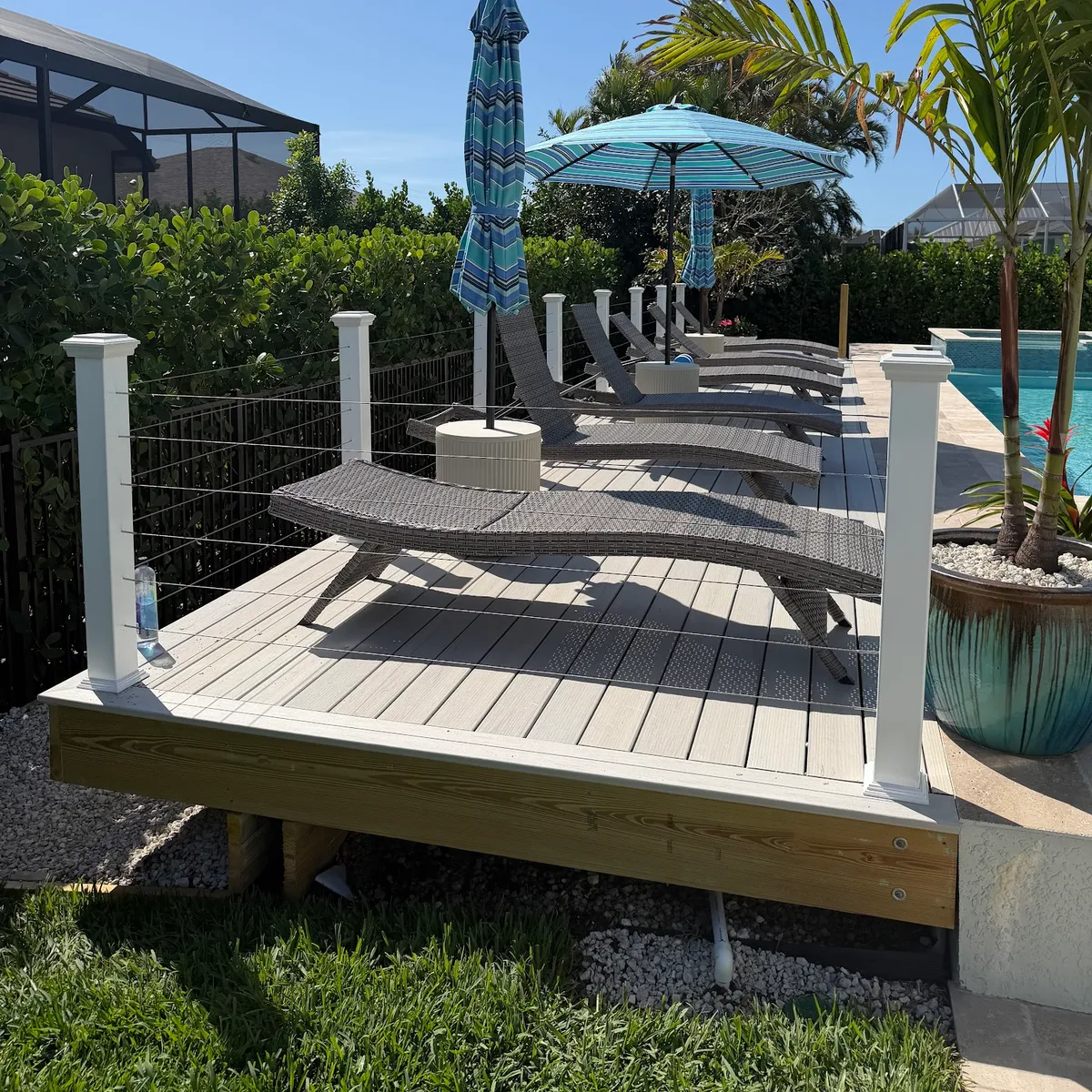 A ground-level composite deck beside a pool, with white posts and cable railing