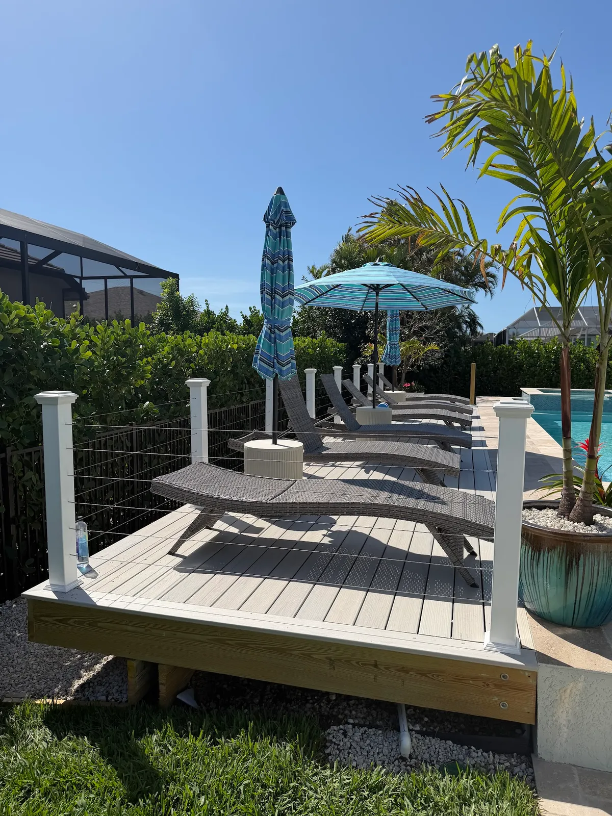 A ground-level composite deck beside a pool, with white posts and cable railing